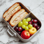 Bright school lunchbox with tuna and cheese sandwiches, apple slices, grapes, and economical healthy foods on a marble background Budget-Friendly School Lunches