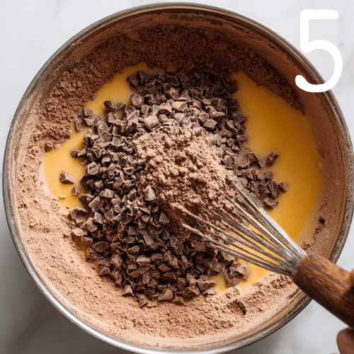 Hand whisking brownie batter in a metal bowl with cocoa powder, melted butter, and chocolate chips on a marble countertop