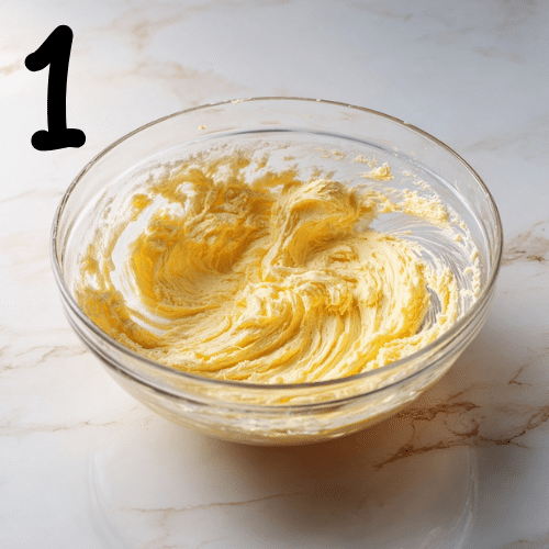 Mixing bowl with butter and sugar creamed until fluffy on a marble countertop, hand mixer resting nearby