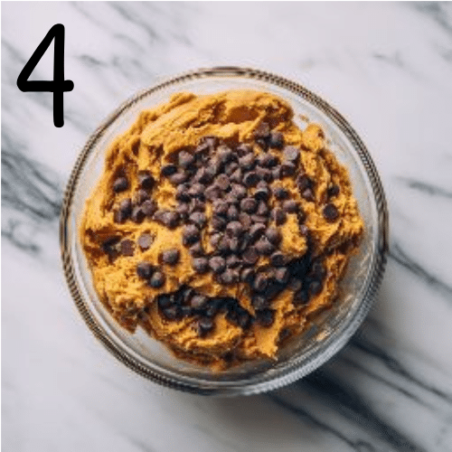 Pumpkin cookie batter in a glass bowl with chocolate chips being stirred in on a marble countertop, cozy fall baking scene