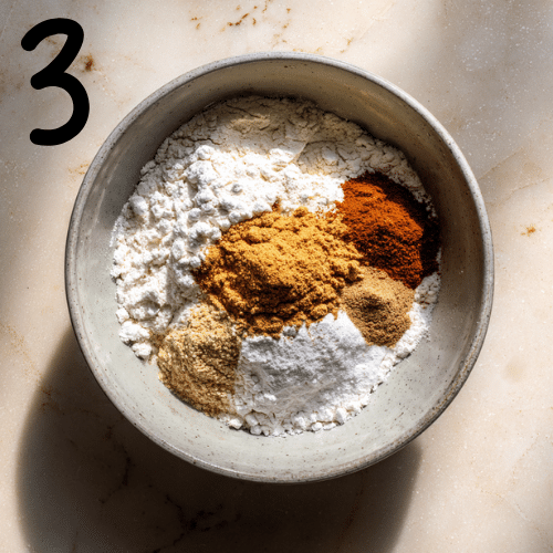 Overhead view of flour, baking powder, baking soda, pumpkin spice, cinnamon, and salt being whisked in a glass bowl on a marble countertop, cozy fall baking scene