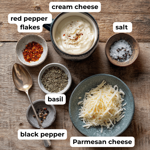 Overhead view of cream cheese, Parmesan, dried basil, red pepper flakes, salt, and pepper on a wooden surface.