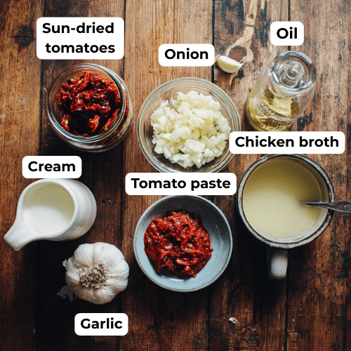 Flat lay of Marry Me Chicken Soup ingredients – sun-dried tomato oil, onion, garlic, tomato paste, chicken broth, and cream on a wooden table.