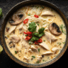 A steaming bowl of Thai Chicken Soup Recipe with sliced chicken, noodles, baby mushrooms, and crushed red peppers in a creamy coconut broth, garnished with coriander and lime.